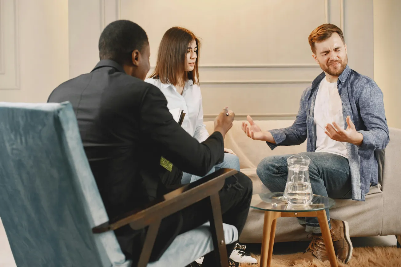 A man in a blue shirt gestures emotionally while talking to a therapist and a woman. The setting is calm with soft lighting, suggesting a therapy session.