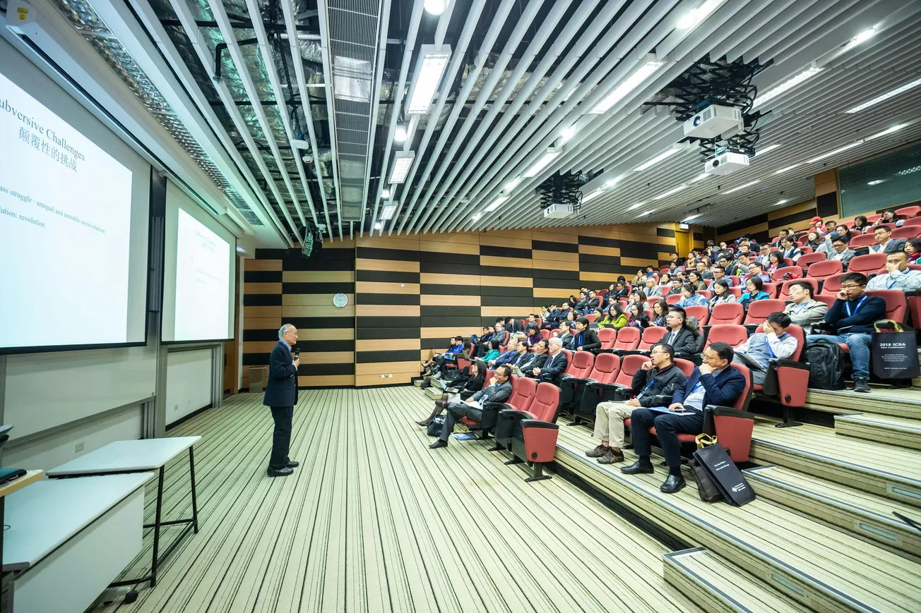 A speaker presents in a modern lecture hall, filled with attentive audience members seated in tiered red chairs. The room has a dynamic, professional atmosphere.