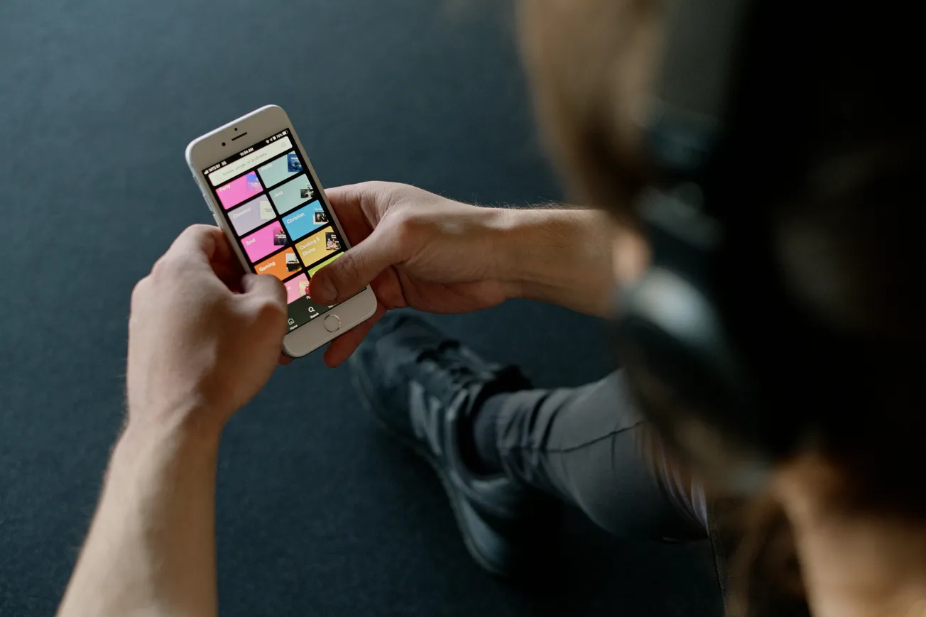A person wearing headphones holds a smartphone displaying a colorful grid of apps, suggesting a relaxed, tech-focused setting on a dark background.