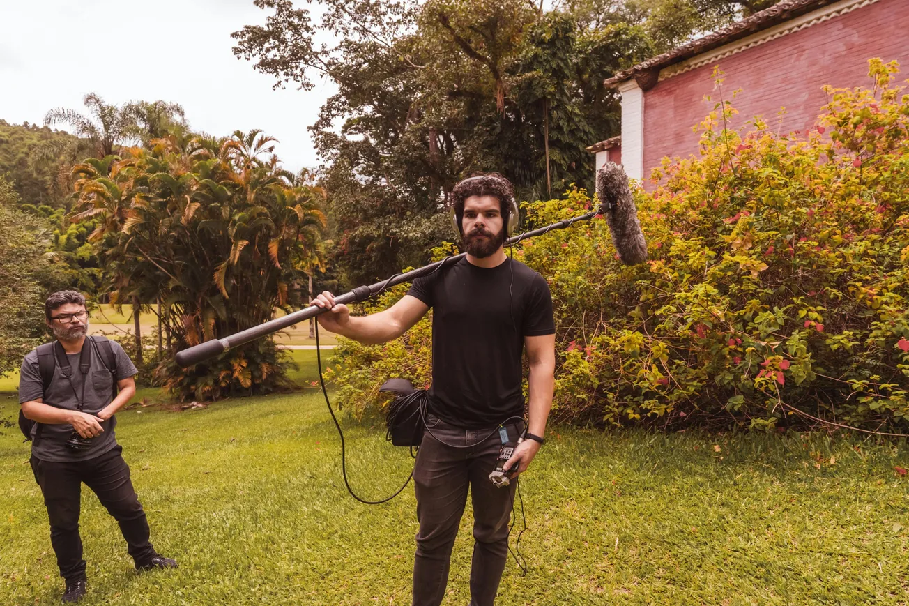 A man holds a boom microphone in a lush garden, wearing headphones and a black t-shirt. Another person stands nearby, both focused on their task.