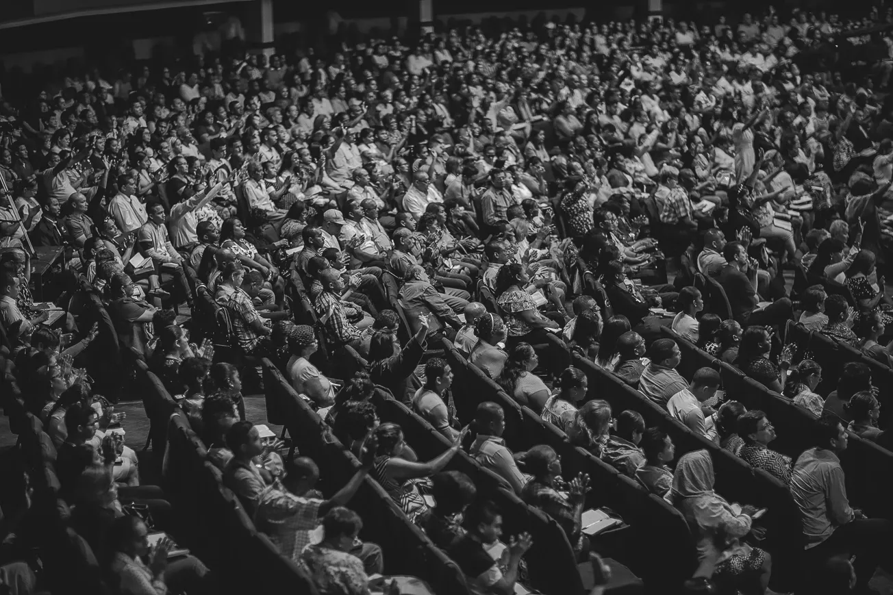 A large, attentive audience fills a theater, captured in black and white. People are seated closely, some clapping, creating a lively, engaged atmosphere.