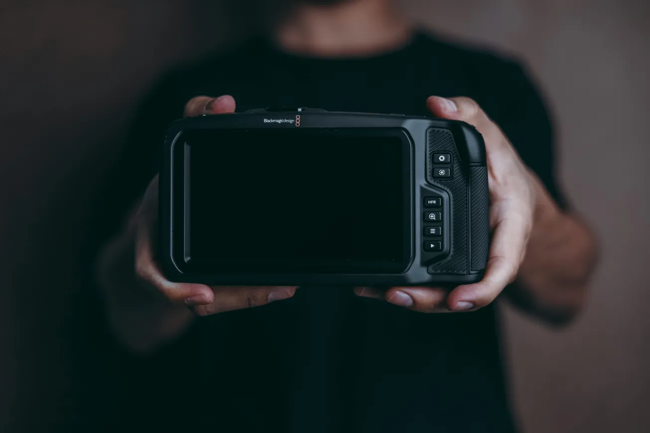 Person holding a large, black camera with a screen facing forward, against a dark background. The mood is focused and professional.