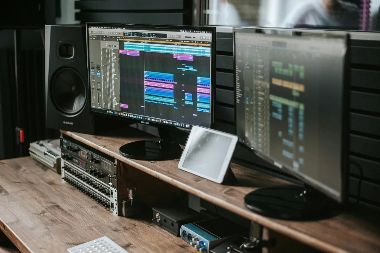 A music production studio setup with two monitors displaying audio editing software, a speaker, and mixing equipment on a wooden desk. A tablet is placed between monitors.