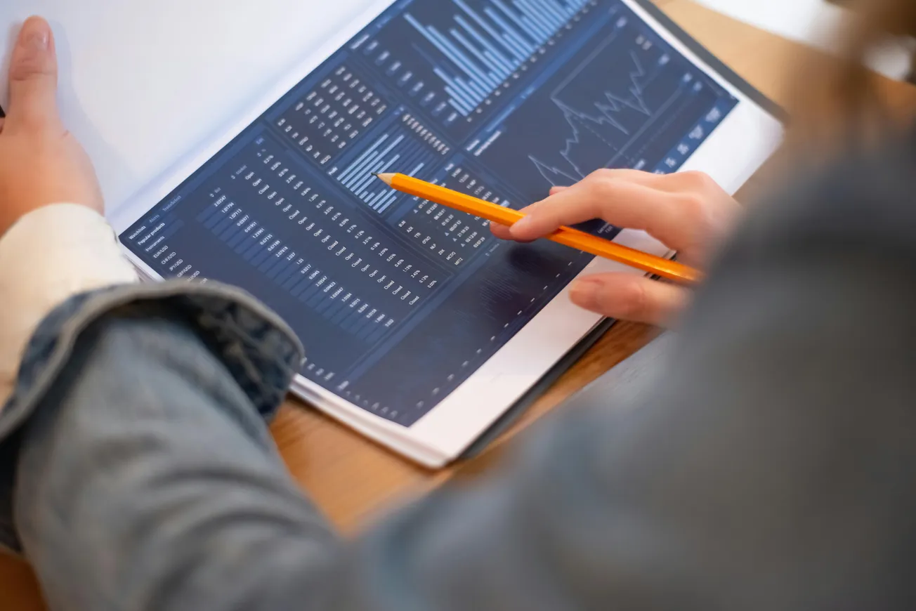Close-up of a person in a denim jacket analyzing graphs and charts with a pencil, conveying focus and analysis in a professional setting.