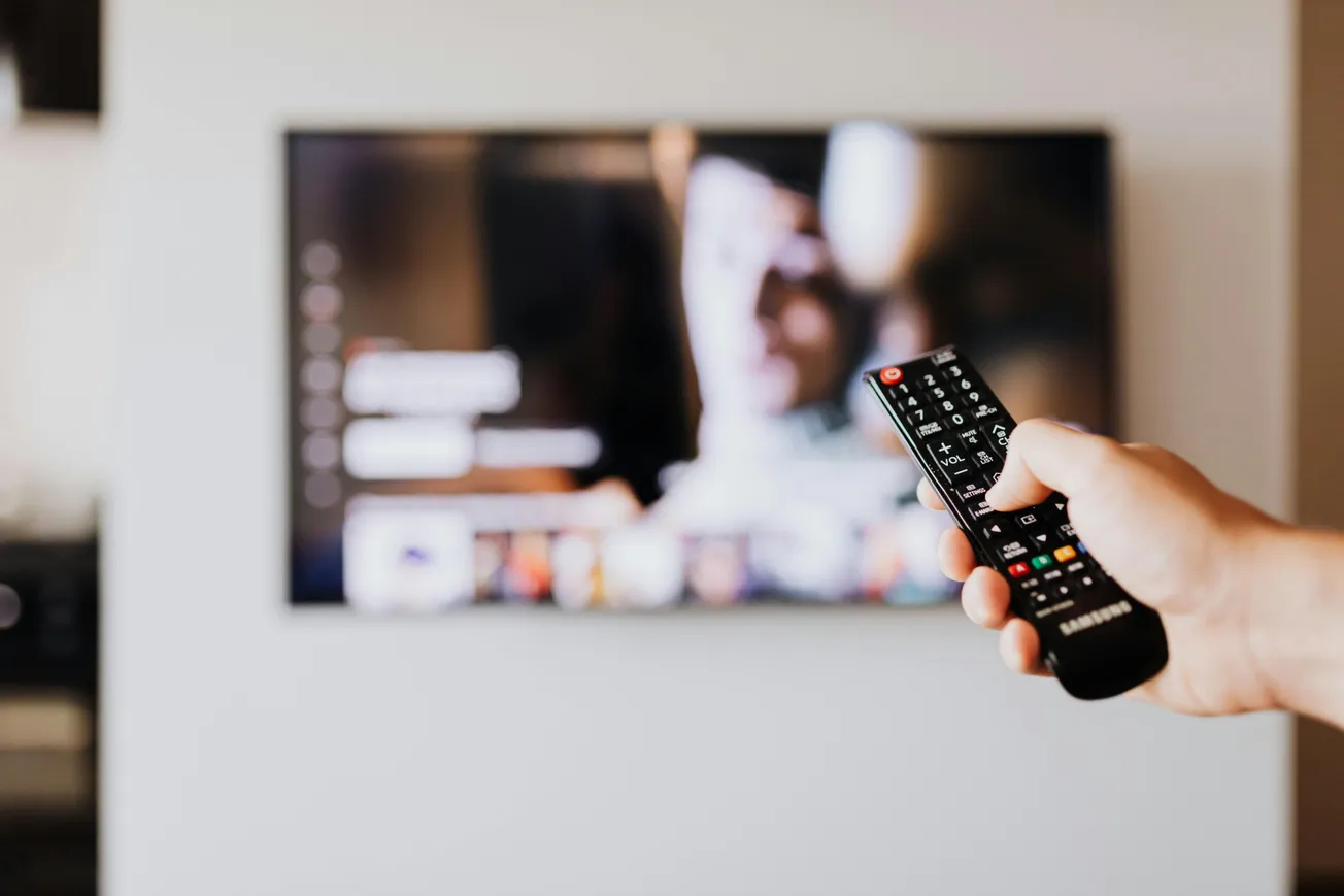 A person holds a Samsung TV remote, pointing it at a blurred TV screen displaying a streaming service interface, conveying a relaxed viewing moment.