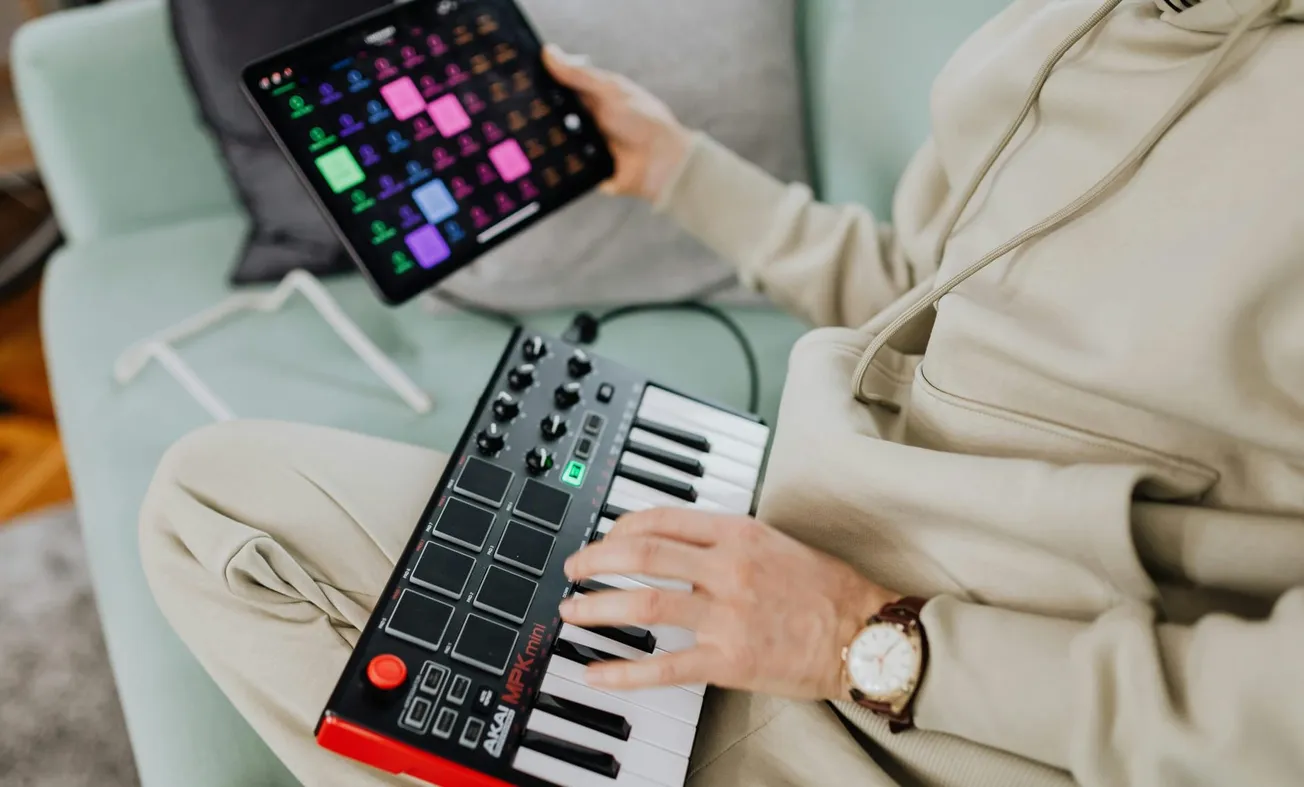 A person in casual clothing sits on a sofa with a MIDI controller on their lap, using one hand on it and the other holding a tablet showing a colorful music app.