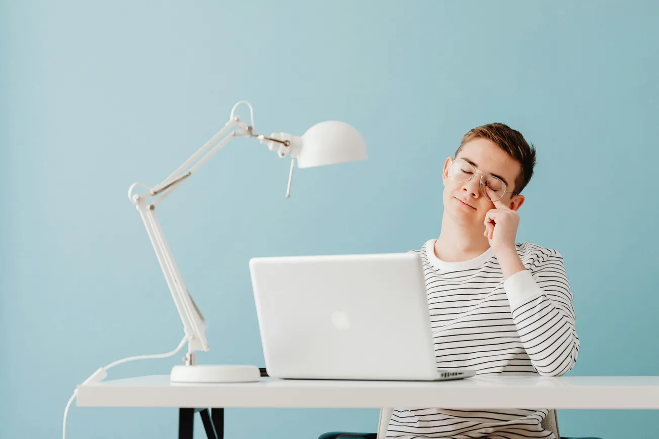 A young person in a striped shirt sits at a desk with a laptop, rubbing their eye. A white lamp is next to them. The mood is tired, set against a blue background.