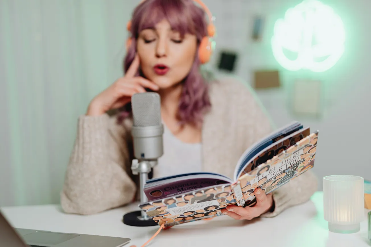 Woman with purple hair and orange headphones speaks into a microphone, reading an illustrated book. The setting is cozy with a soft, glowing light.