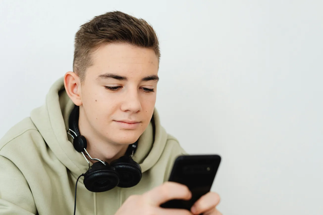 Young man in a green hoodie with headphones around his neck looks at a smartphone, conveying a focused and relaxed expression against a plain white background.
