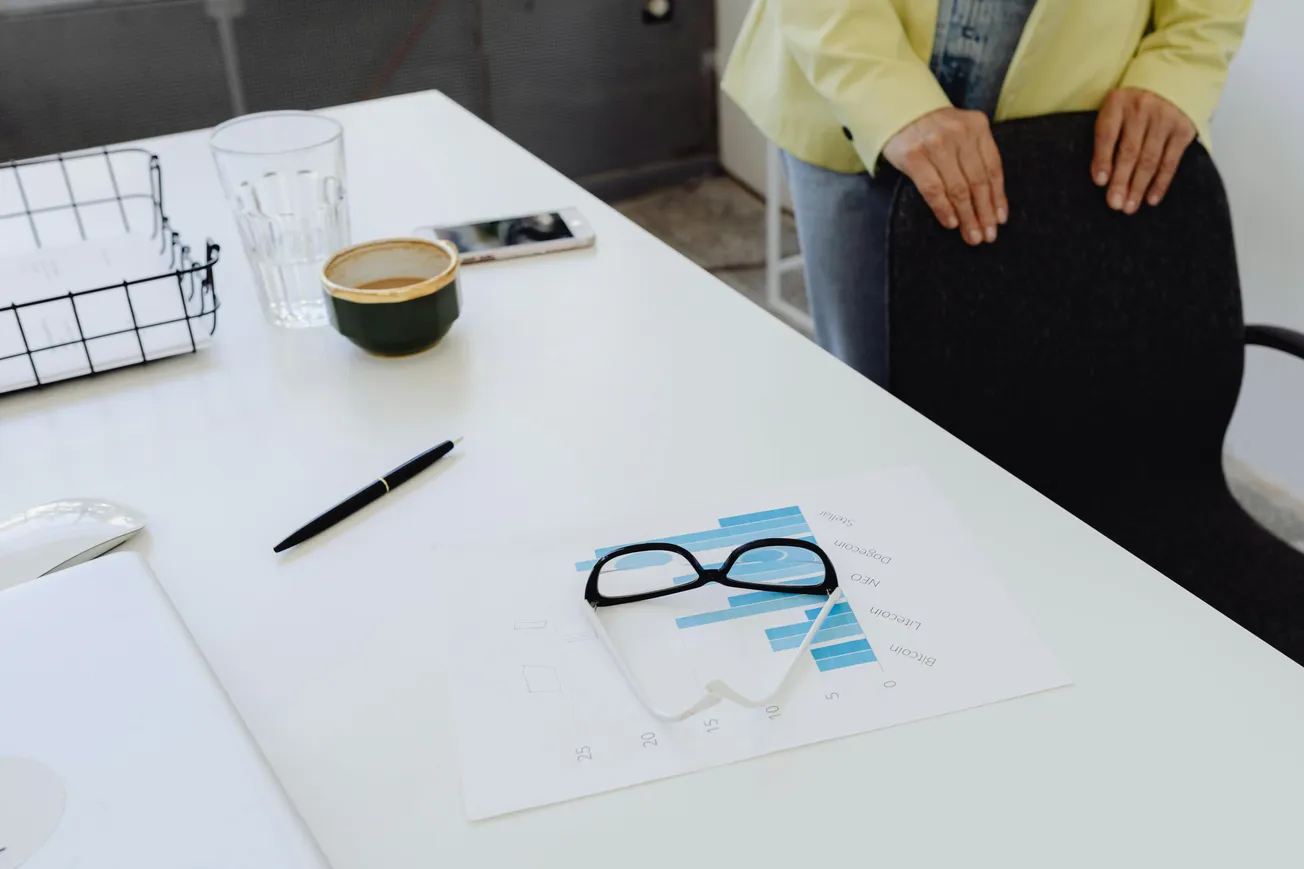 A desk with charts, glasses, a pen, phone, and drink show a workspace vibe. A person stands beside with hands on a chair, creating a calm, organized tone.