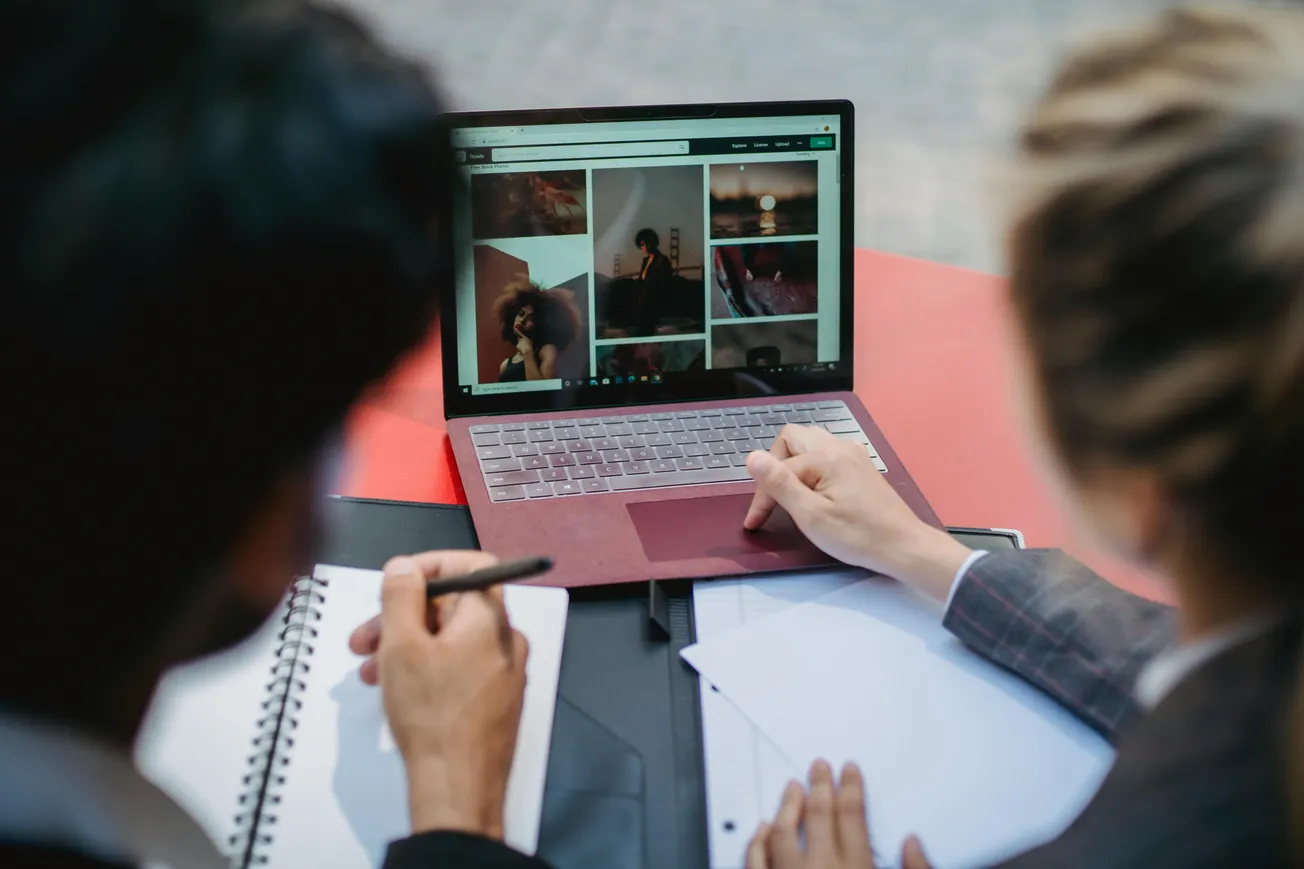 Two people working together, viewing images on a laptop. One is writing in a notebook, creating a collaborative and focused atmosphere.