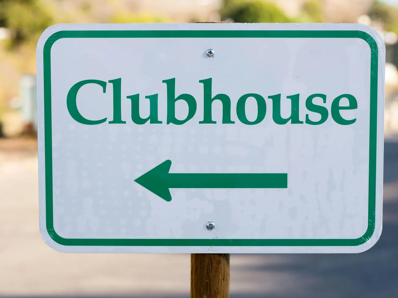 A green and white sign with the word "Clubhouse" and a left-pointing arrow, indicating direction. The sign is mounted on a wooden post outdoors.