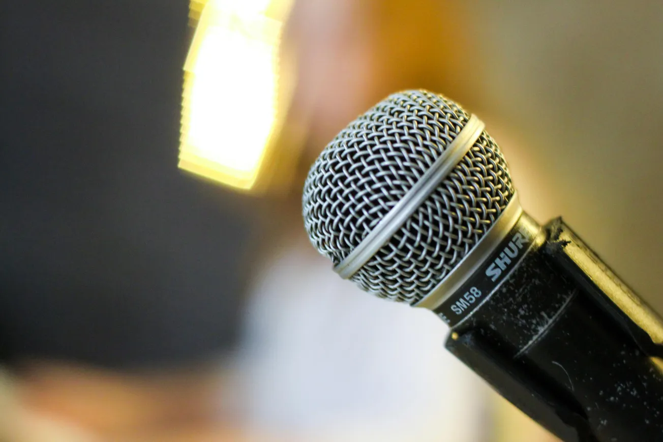 Close-up of a silver Shure SM58 microphone with a mesh grille, slightly tilted, set against a blurred background with warm lighting, suggesting a lively performance atmosphere.