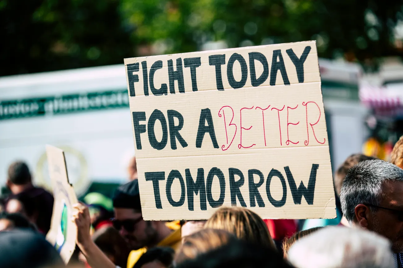 Protest scene with a crowd holding a sign reading “Fight today for a better tomorrow.” Keywords: protest, sign, crowd, change, hope, determination.