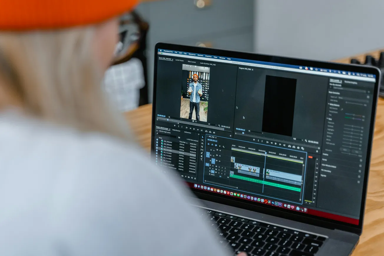 A person in a beanie edits a video on a laptop using Adobe Premiere Pro. The screen displays video clips and editing timelines, suggesting a focused work environment.