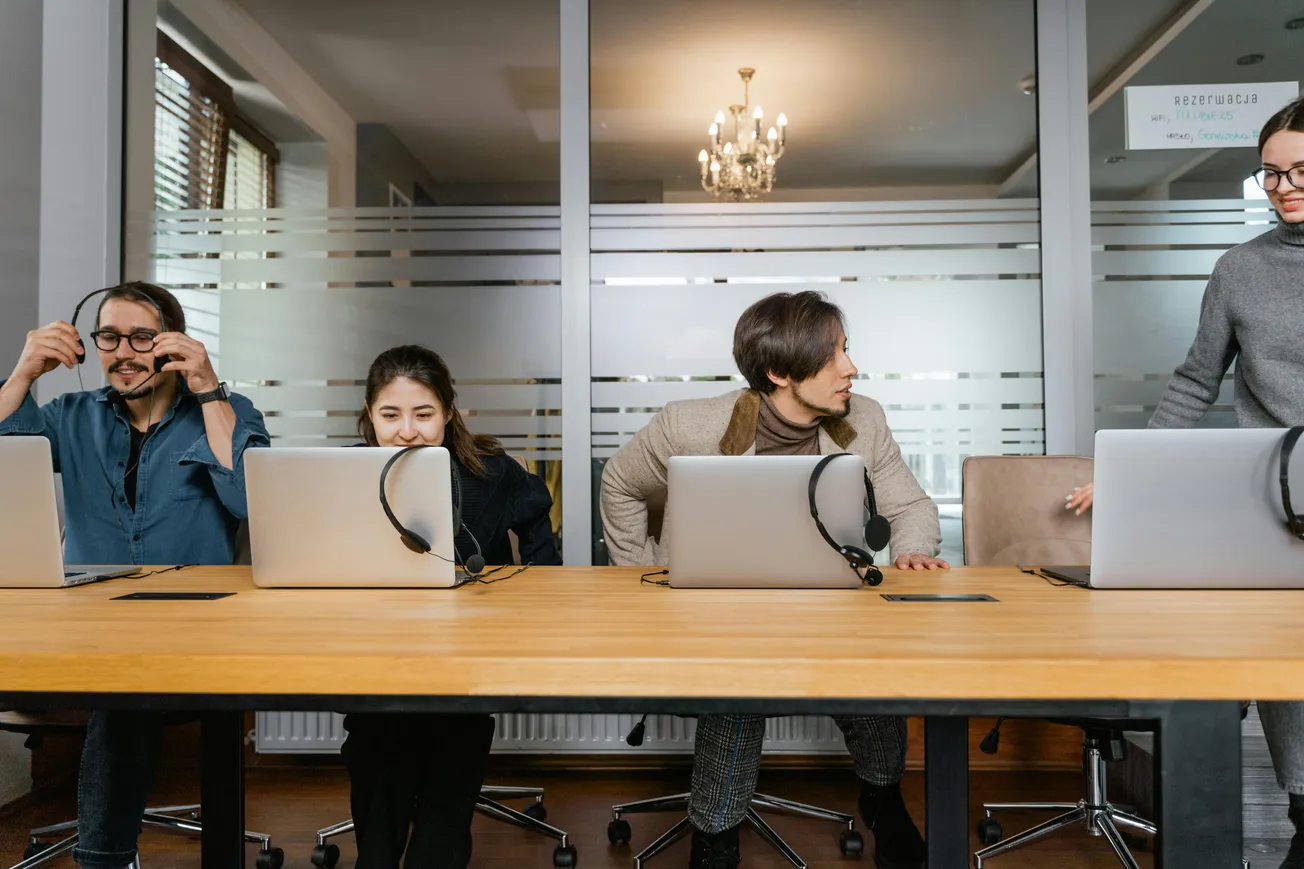 Four people in an office sit at a table with laptops and headsets. One is adjusting their headset. They appear focused and engaged in conversation.