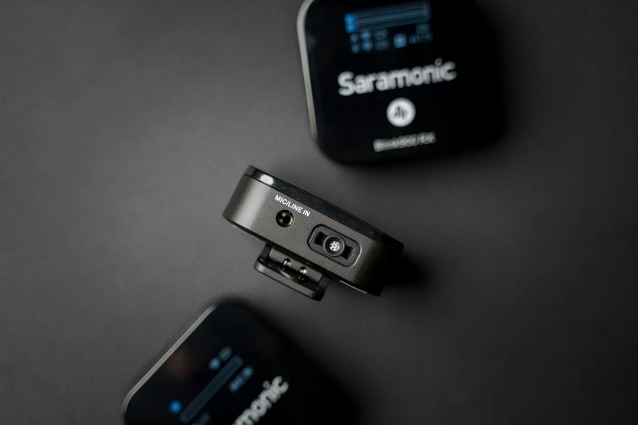 Close-up of a black Saramonic wireless microphone system on a dark surface. The central unit shows inputs and buttons, conveying a sleek, modern design.