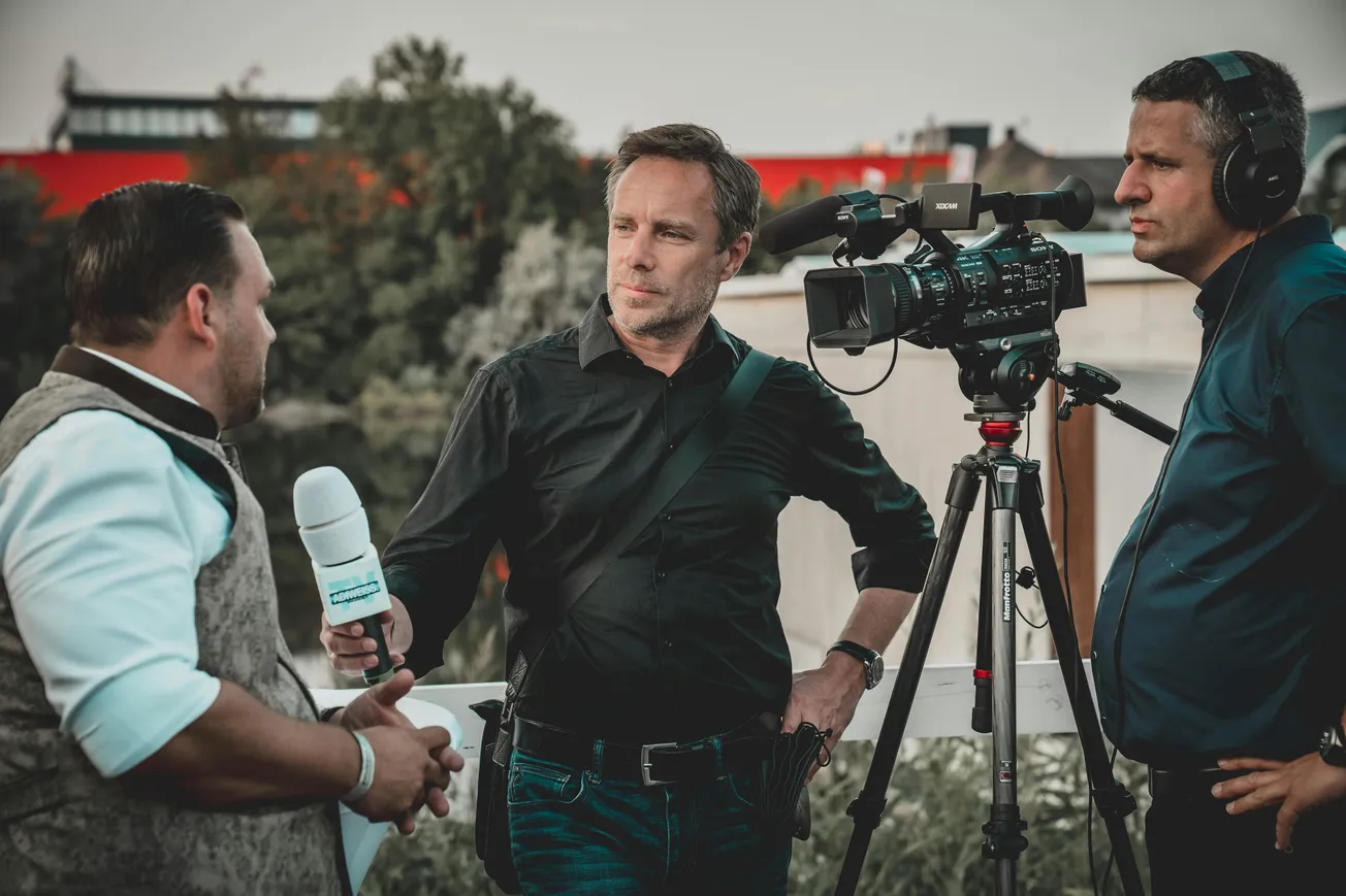 A cameraman films an outdoor interview between a reporter holding a microphone and a man in a vest. The scene conveys focus and professionalism.