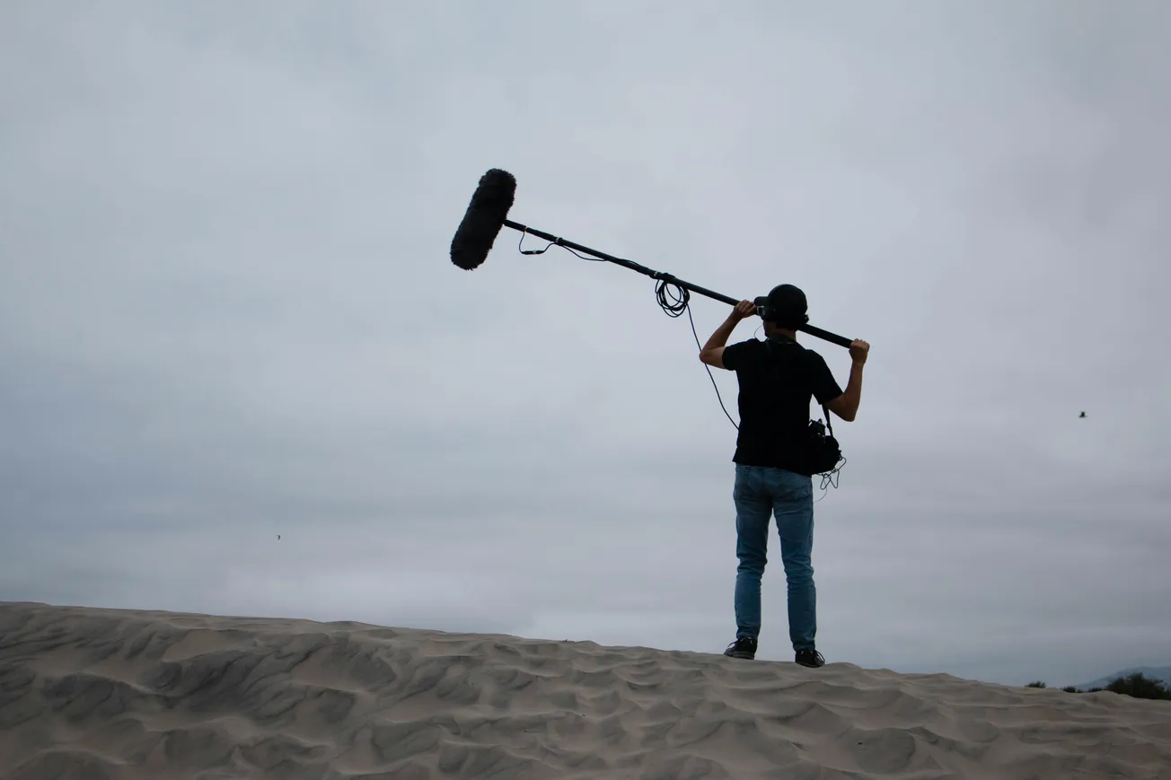 Person stands on sand dune holding a boom mic against a cloudy sky, capturing sound. Atmosphere is calm and focused, with muted tones.