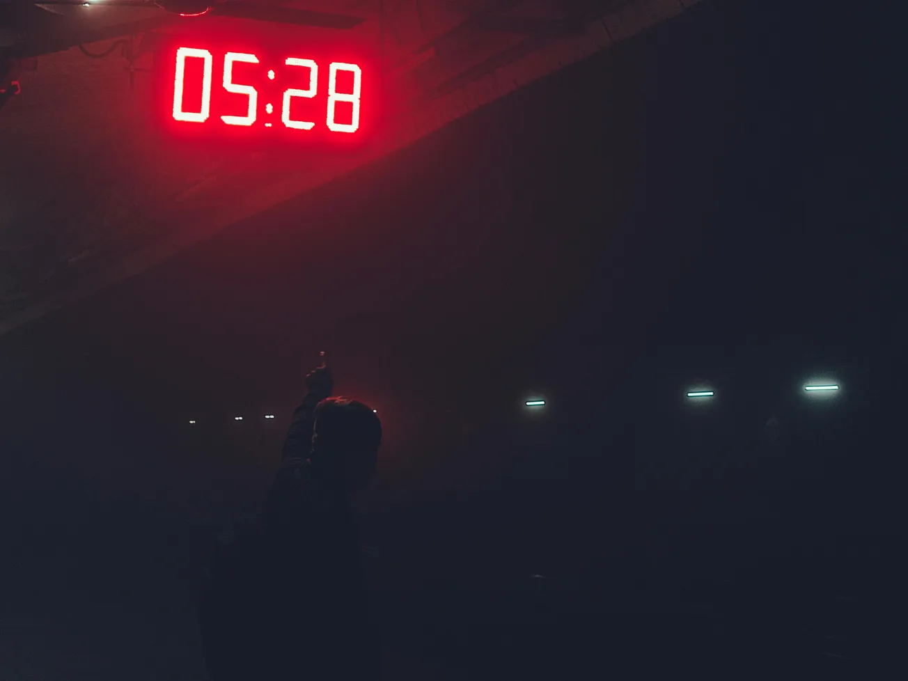 A person in a dark space points upward toward a glowing red digital clock displaying "05:28." The scene feels mysterious and somber.