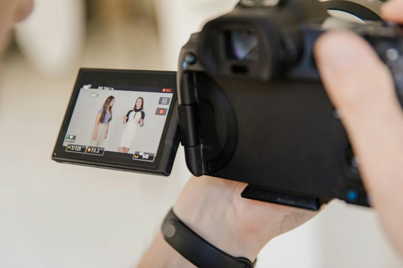 Close-up of a camera screen displaying two women standing side by side, dressed in light clothing, being recorded. The focus is on the camera's perspective.