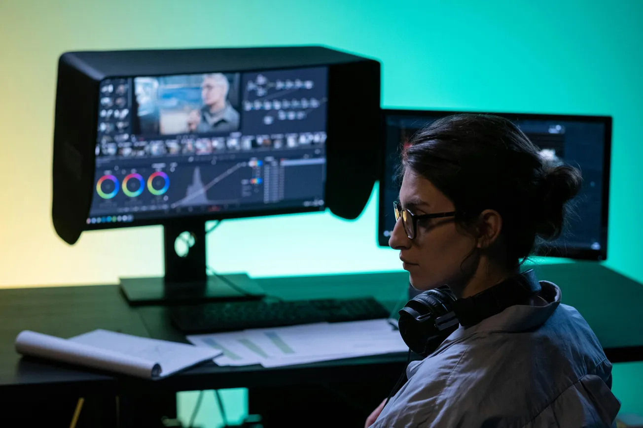 A person with glasses and headphones sits at a desk editing video on dual monitors against a green and yellow backdrop. The atmosphere is focused.