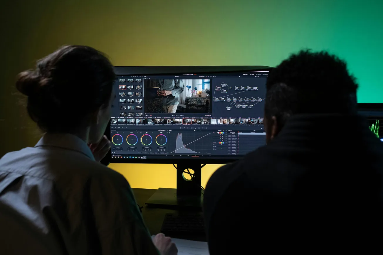 Two people sit at a desk editing video on a large dual-monitor setup, with graphs and footage visible, against a green-yellow gradient wall.
