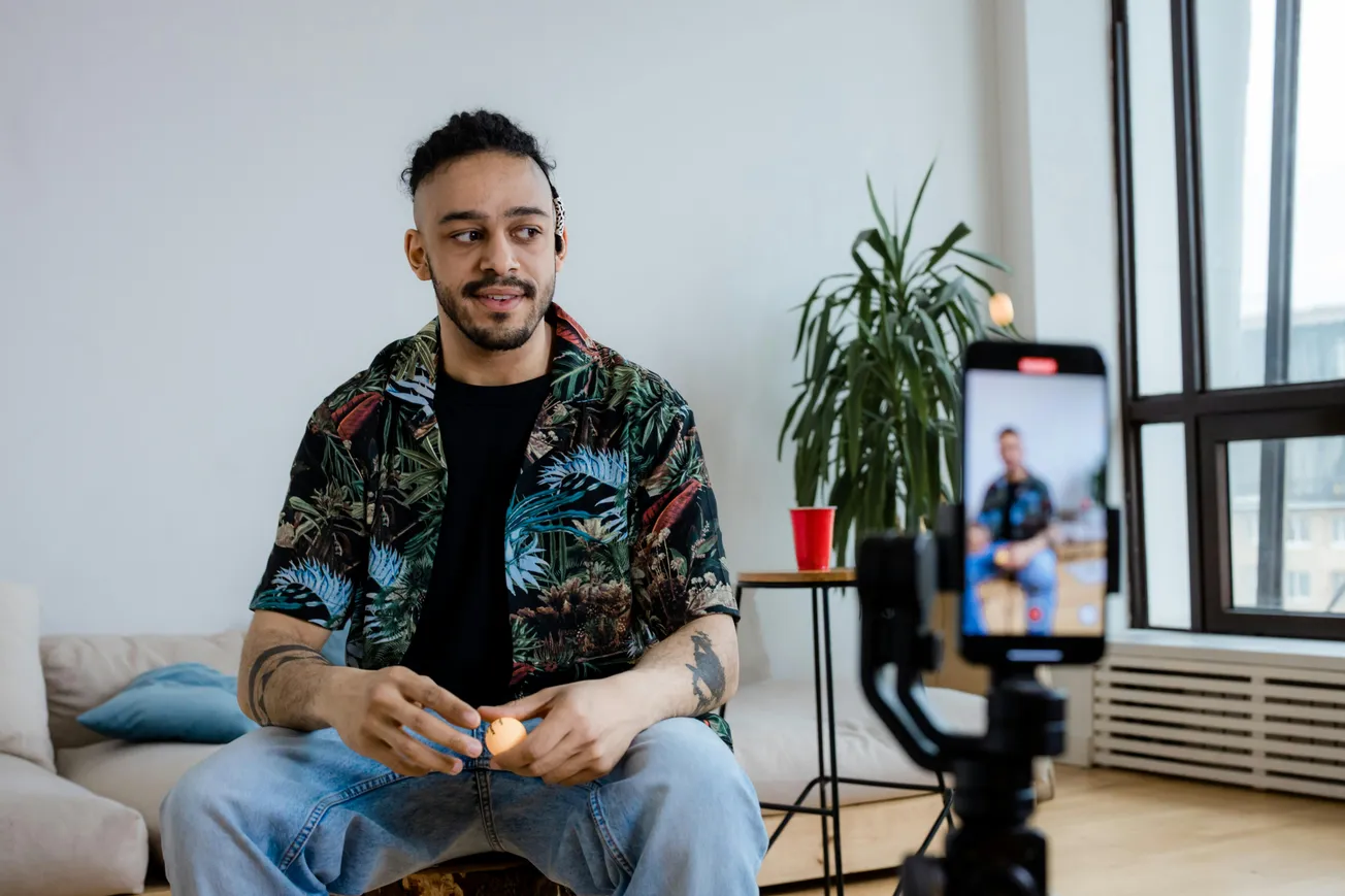 A man in a tropical shirt sits casually on a couch, filming himself with a smartphone on a tripod. The room is bright, with a plant and red cup nearby.