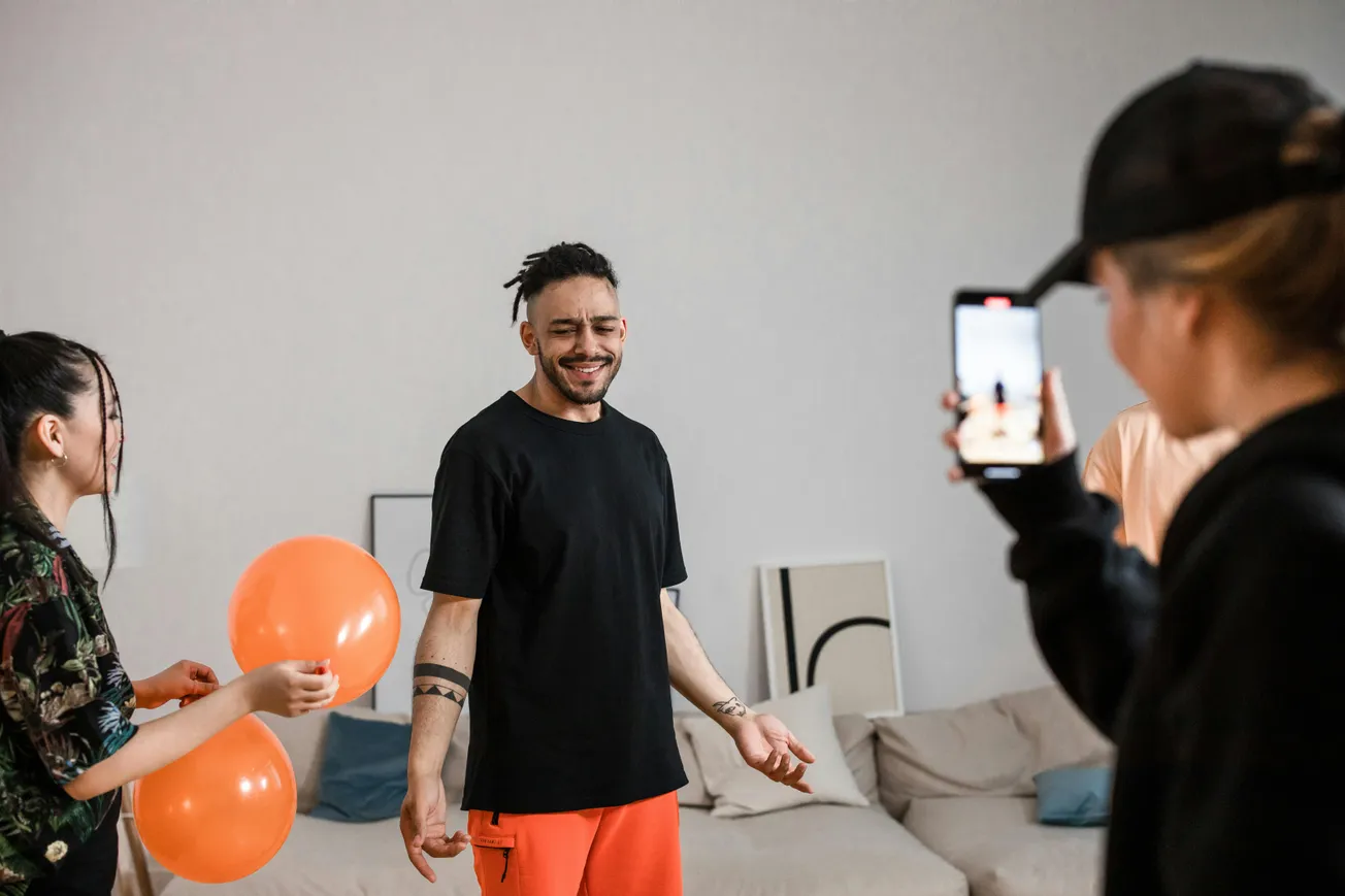 Three people in a living room: one holding orange balloons, another in orange pants smiling with eyes closed, and a third taking a photo with a phone.