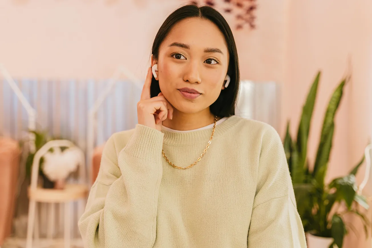 A woman in a cozy, light-colored sweater touches a wireless earbud. She appears thoughtful, in a softly lit room with plants and modern decor.