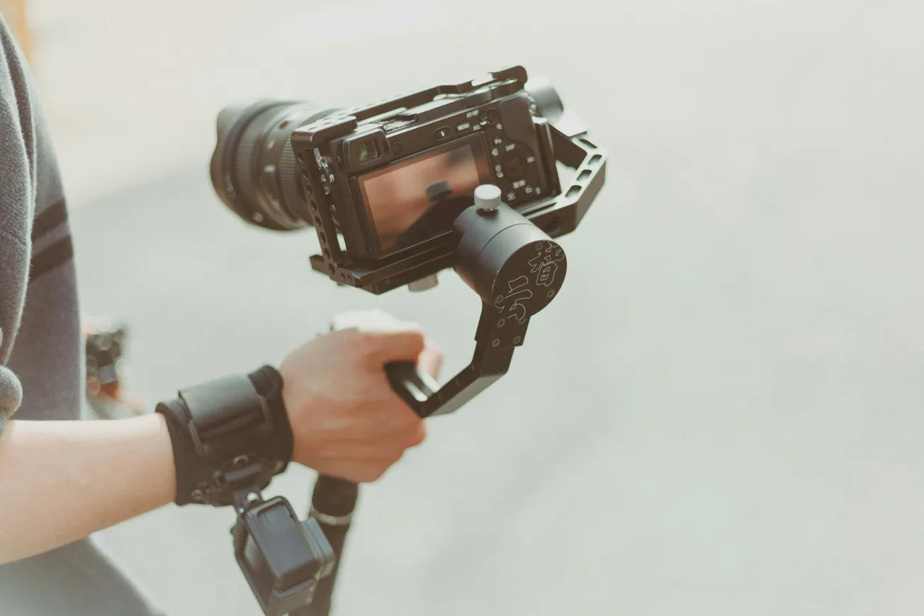 Close-up of a person's hands holding a DSLR camera mounted on a gimbal. The scene is well-lit, conveying a sense of focus and professionalism.