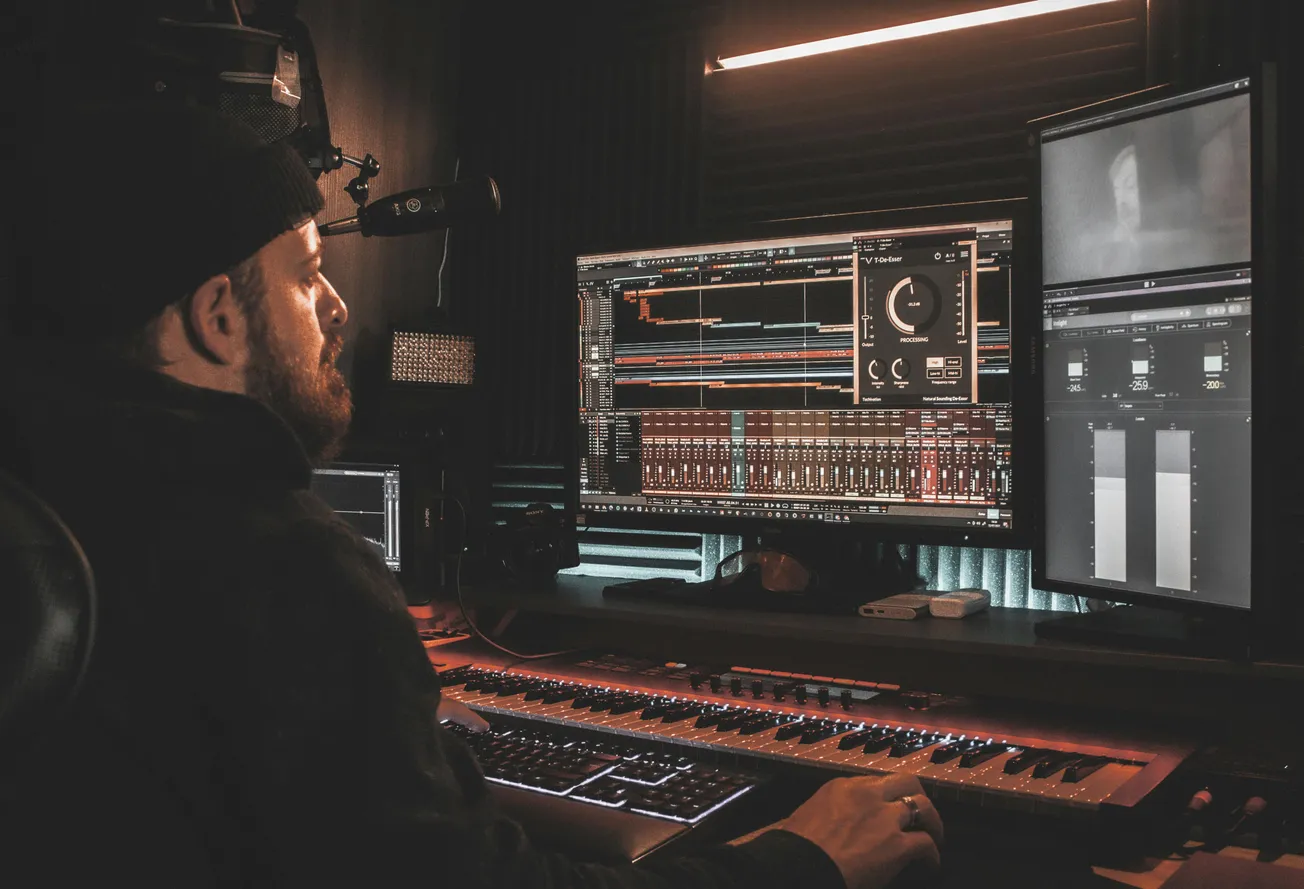 A person wearing a beanie is working in a dimly lit sound studio, focused on two large monitors displaying complex audio editing software.