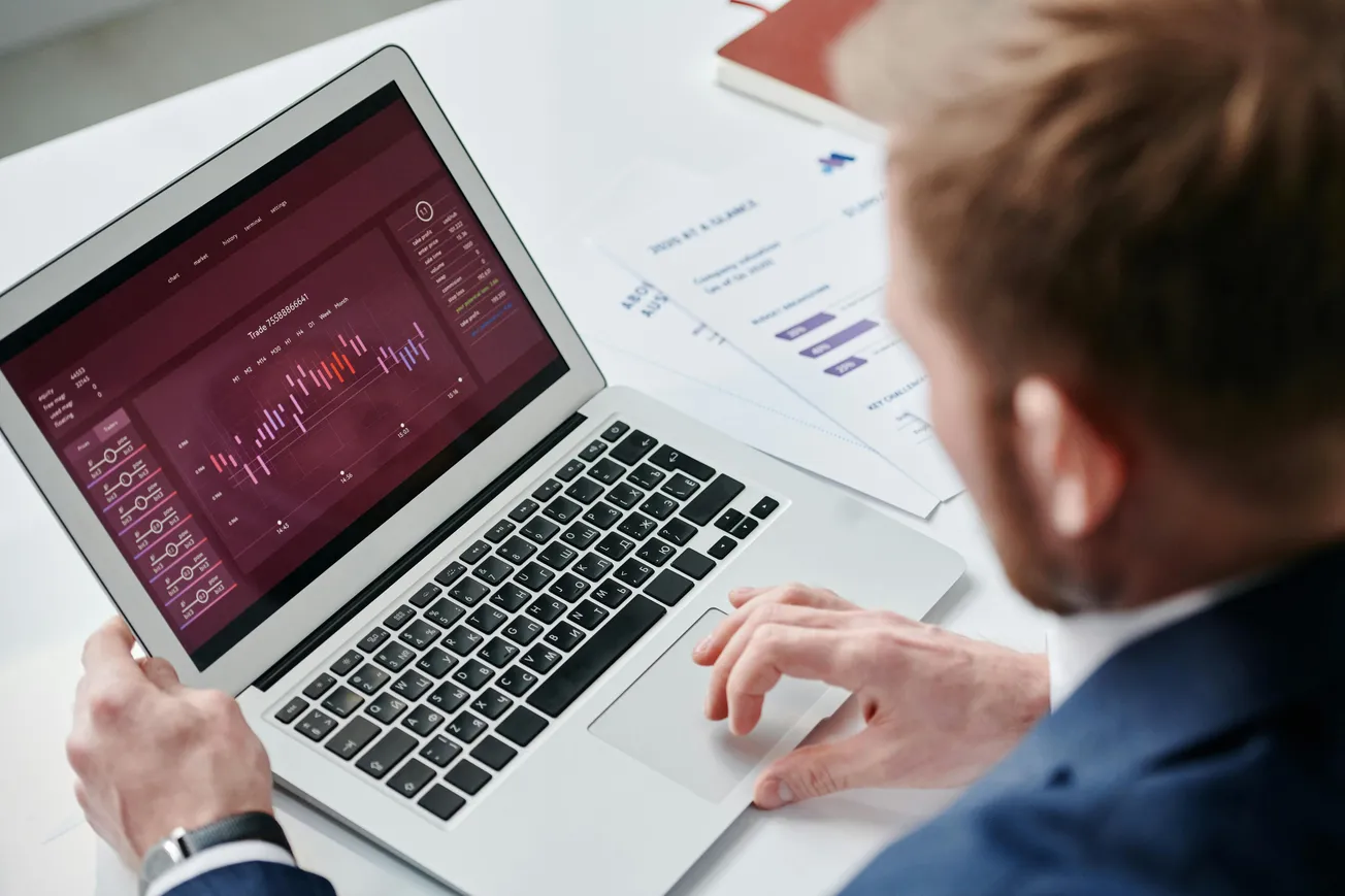 A person in a suit is using a laptop displaying colorful financial graphs and charts. Papers and a pen are on the desk, indicating a business setting.