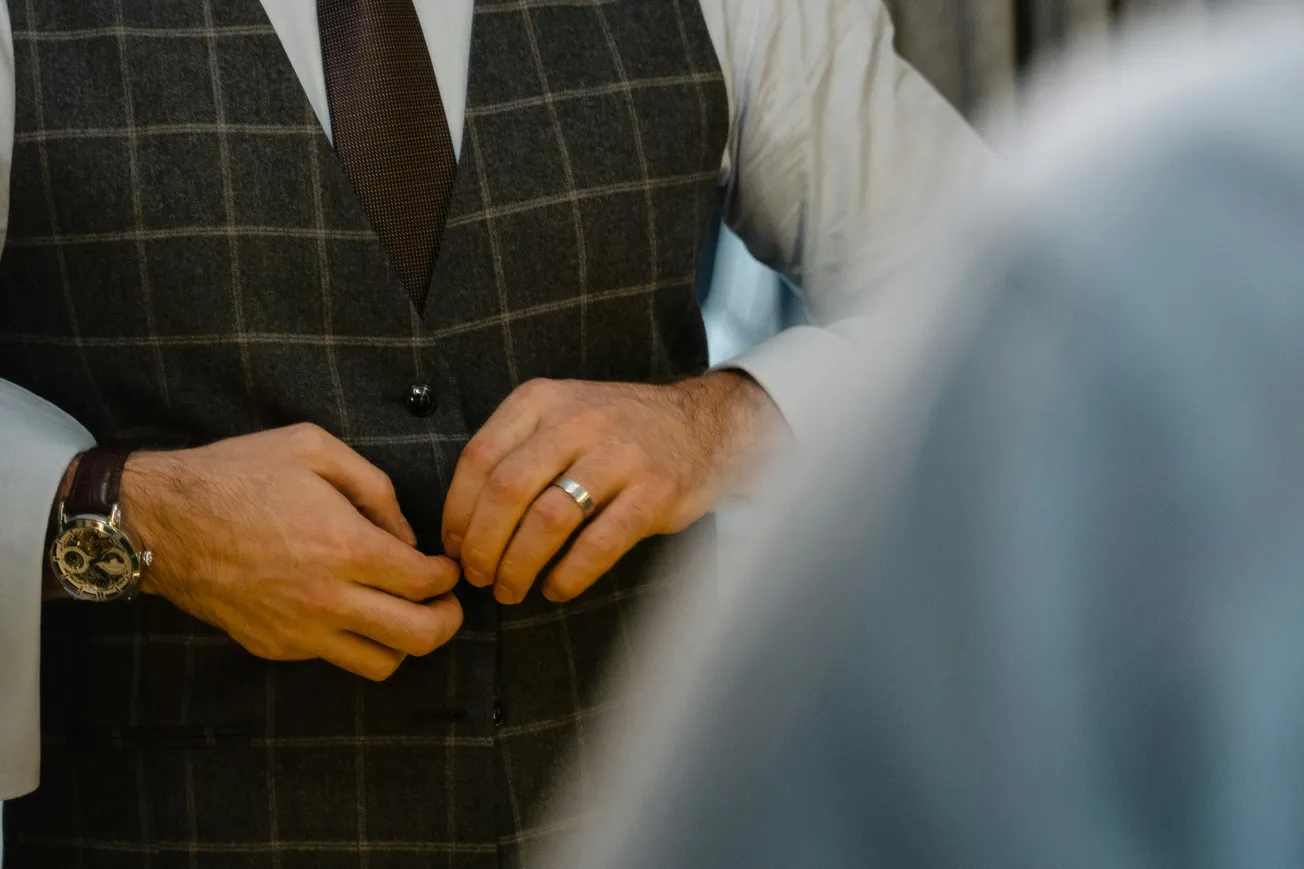 A man adjusts his gray plaid suit vest, showcasing a brown tie and a silver wristwatch. The image conveys a sense of elegance and preparation.