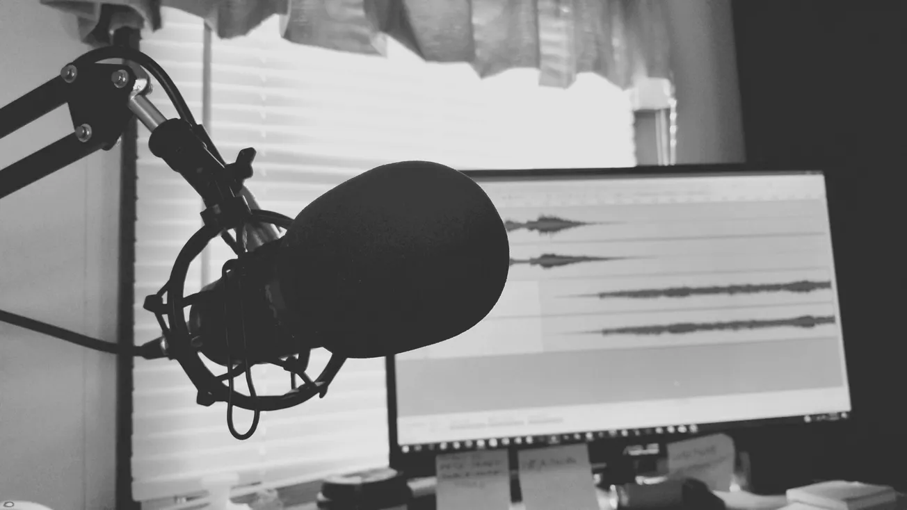 Black and white image of a podcast setup featuring a microphone on an arm stand in the foreground and an audio waveform on a computer screen in the background.