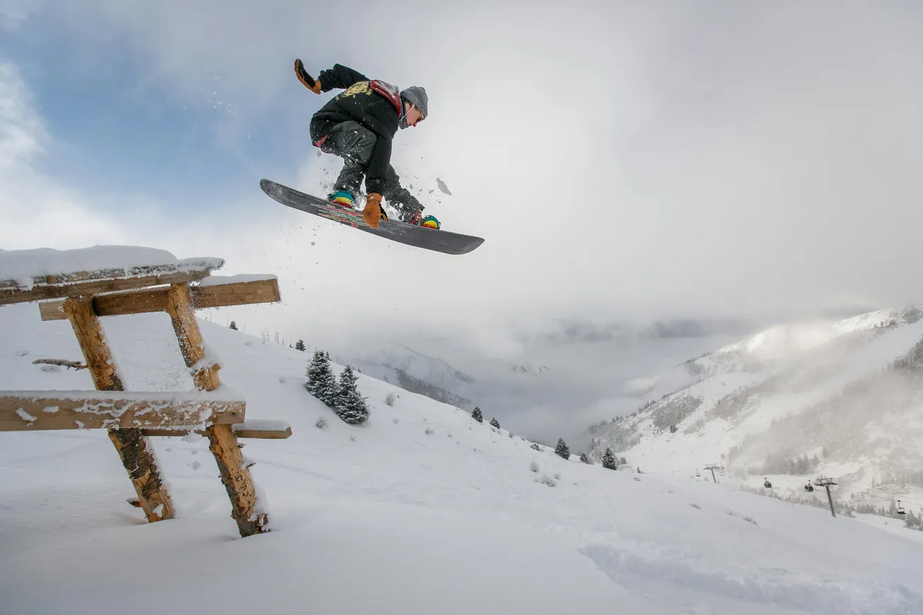 Snowboarder in mid-air, leaping off a snow-covered picnic table on a mountain slope. The background shows snowy peaks under an overcast sky.