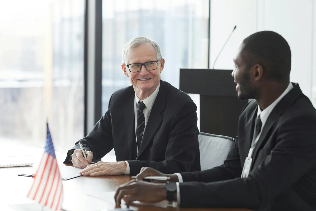 Two men in suits sit at a conference table, engaged in conversation; an American flag is visible. The atmosphere is professional and collaborative.