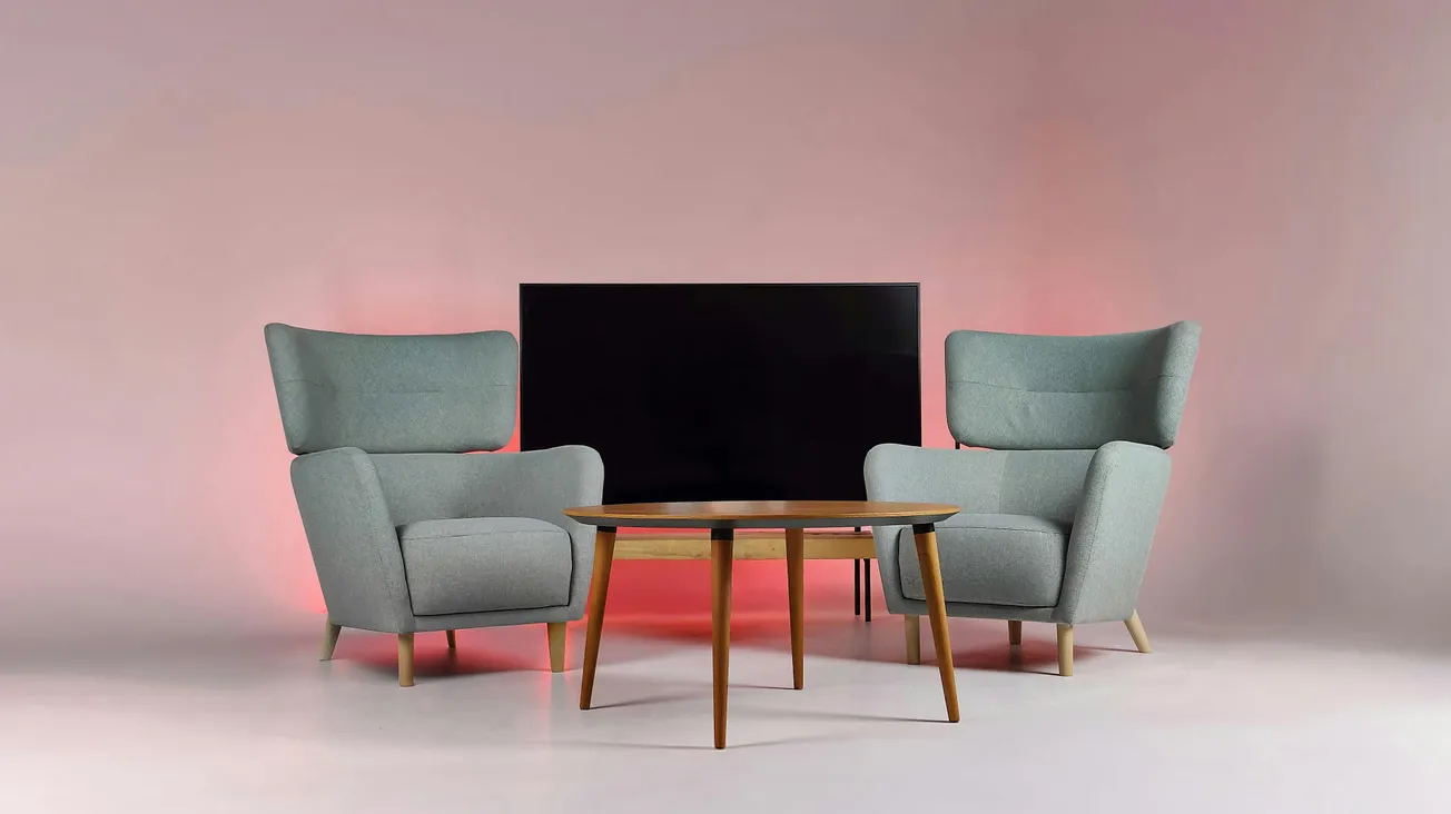 Two gray armchairs flank a wooden coffee table in front of a black TV screen. The background is softly lit with a warm pink glow, creating a cozy ambiance.