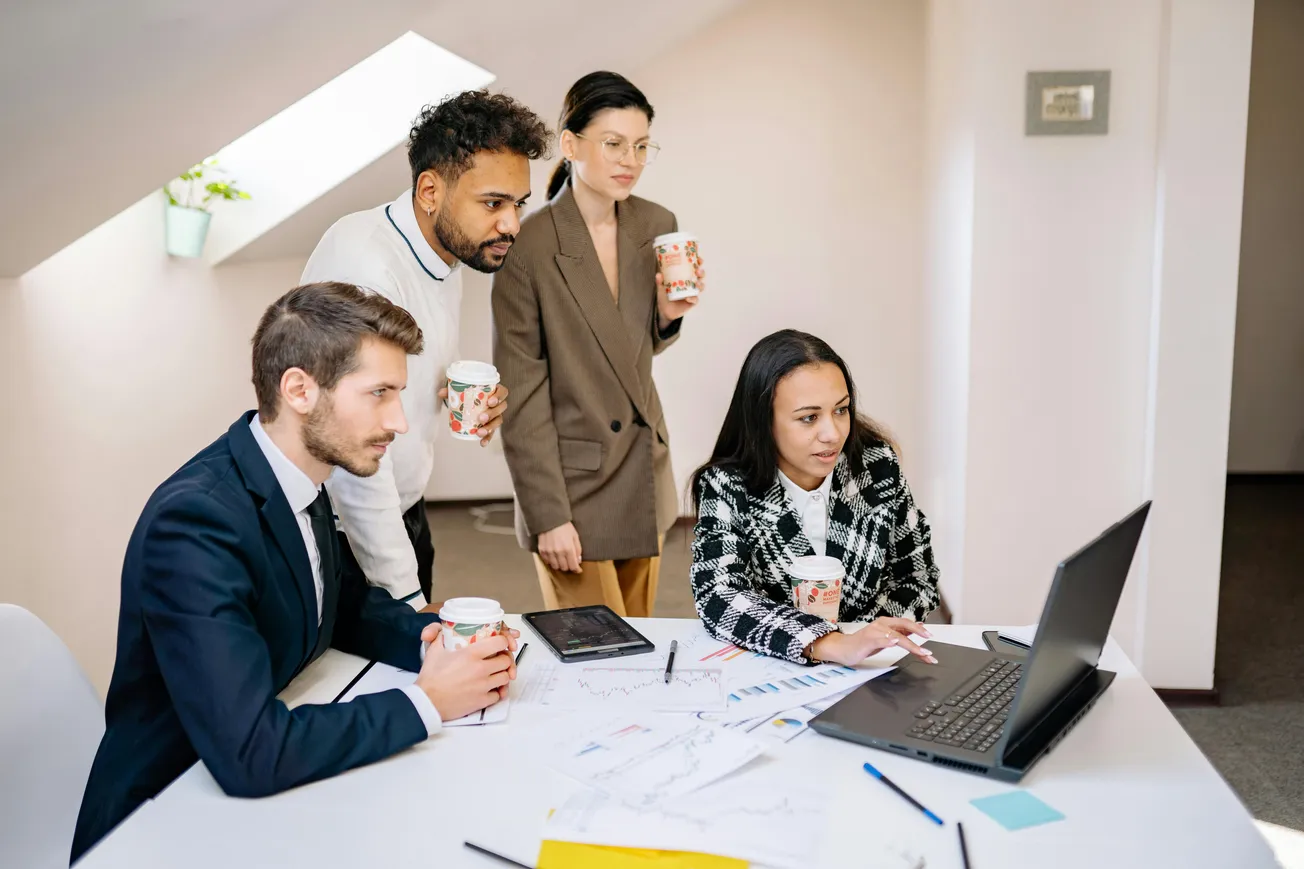 Four colleagues, two men and two women, gather around a laptop, focused and holding coffee cups. Documents with charts lie on the table. Business meeting.