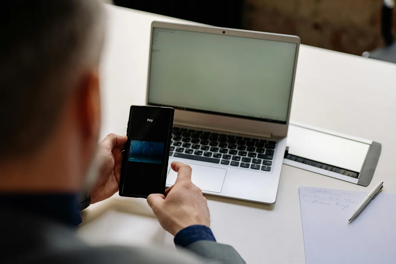 A person interacts with a smartphone showing a payment app, seated at a white desk with an open laptop, papers, and a pen, suggesting a work setting.