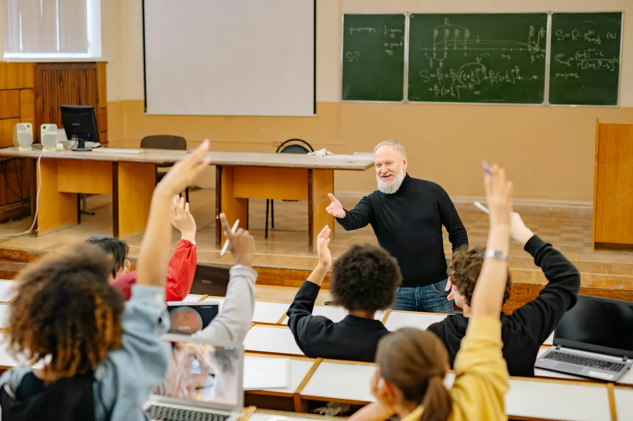 A bearded professor in a black sweater engages with eager students raising hands in a classroom. Chalkboards display complex equations, creating an energetic, academic atmosphere.