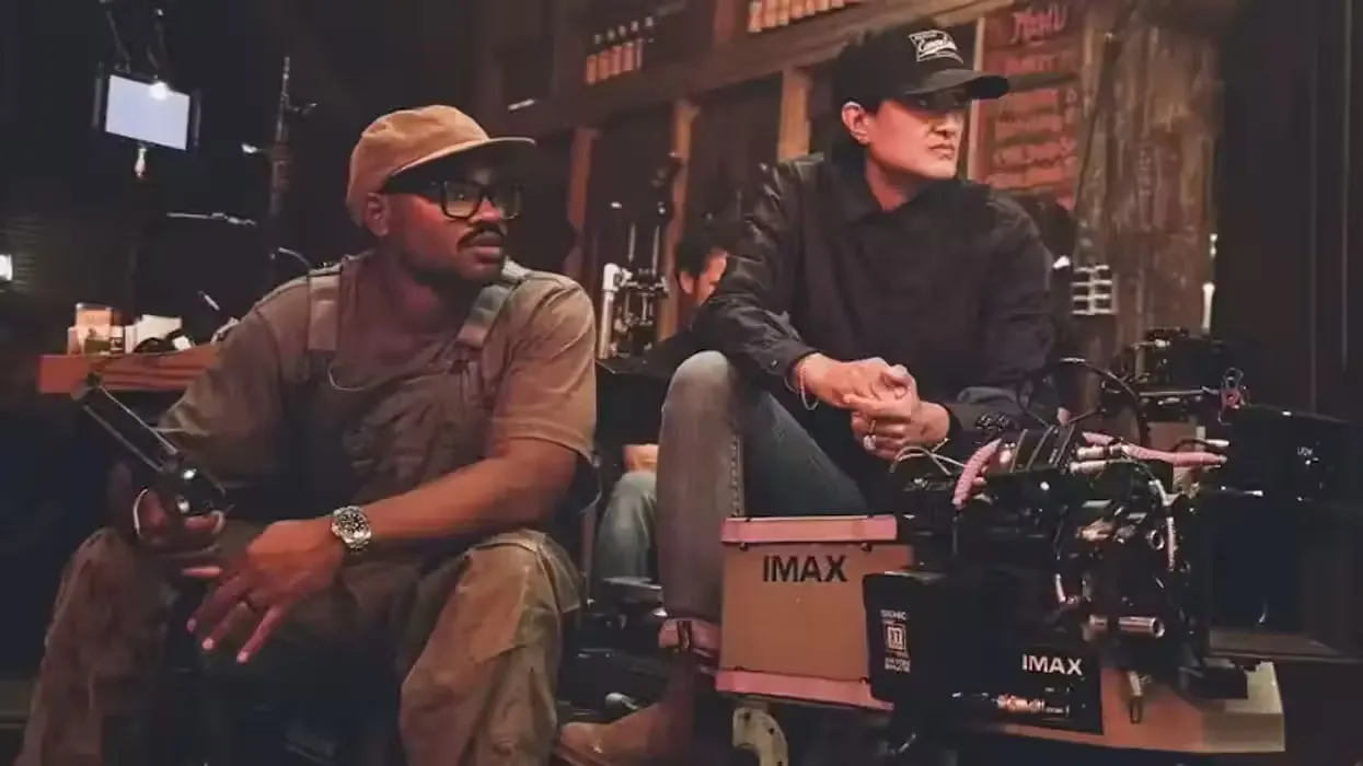 Two filmmakers sit on set beside an IMAX camera. The scene is dimly lit, conveying focus and concentration. One holds a remote, eyes on the scene.