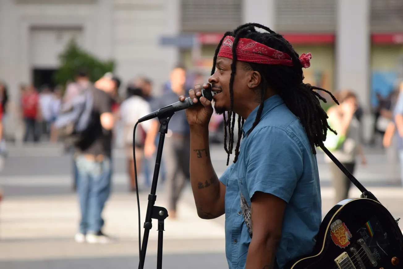 Musician with dreadlocks and red bandana sings passionately into a microphone on a city street. A guitar is slung over their shoulder. Passersby in the background.