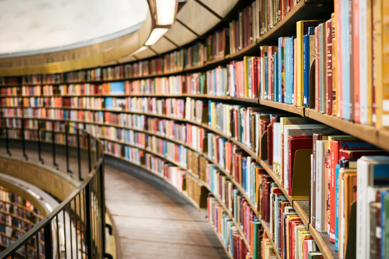 Curved library bookshelves filled with colorful books, creating a cozy, inviting atmosphere. Warm lighting enhances the serene, academic setting.