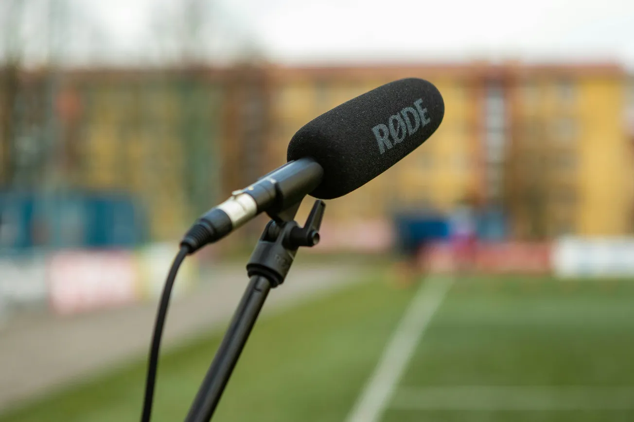 A RODE microphone on a stand is positioned outdoors, with a blurred sports field and buildings in the background. The setting conveys a professional audio setup.