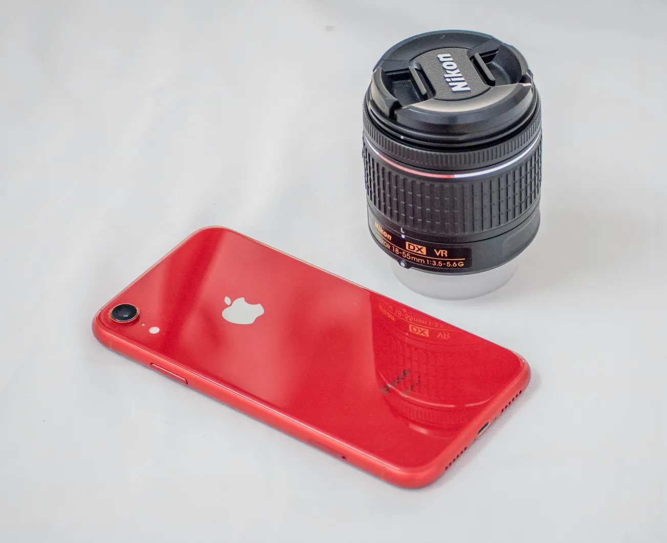 A red smartphone with a white logo lies beside a black Nikon camera lens on a white surface. The scene conveys a tech-savvy and creative vibe.
