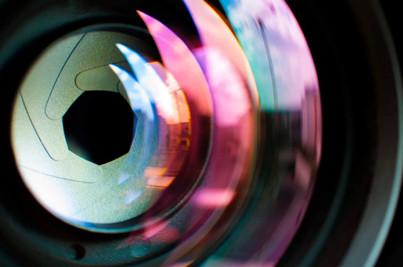 Close-up of a camera lens with colorful reflections. The aperture blades are visible, creating a dynamic and vibrant abstract look, evoking creativity.
