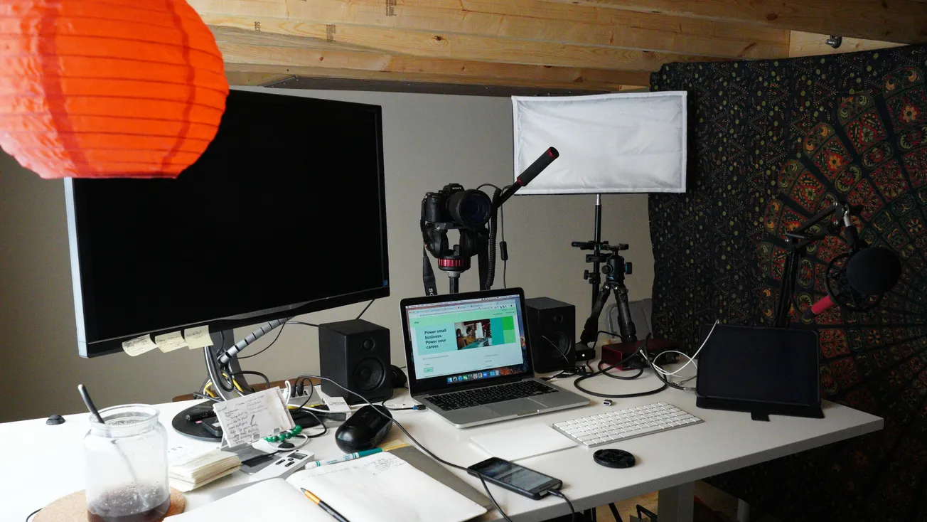 A home recording studio setup with a laptop, large monitor, microphones, and lighting. Cozy and cluttered, with an orange lantern adding warmth.