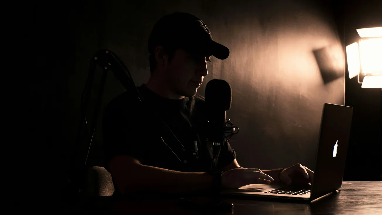 A person in a dim room, wearing a cap, types on a laptop. A microphone and soft light create a focused, contemplative atmosphere.