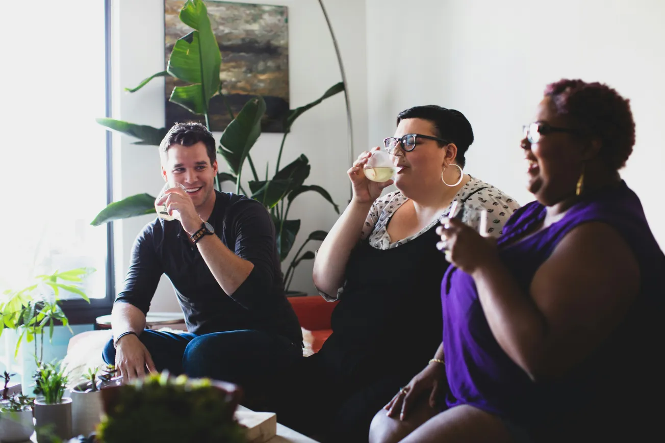 A group of three people sitting and laughing together indoors, drinking from cups. The scene is relaxed with plants in the background and a bright, cheerful atmosphere.